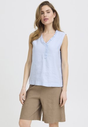 Kvinde iført ærmeløs lyseblå top med flæser og V-hals samt løse brune knælange shorts, stående mod en ensfarvet lys baggrund.