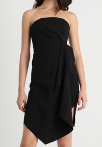 Černé šaty bez ramínek s přiléhavým živůtkem, římským postranním detailem a asymetrickým lemem. Vyrobeno z hladké tkaniny s matným povrchem.