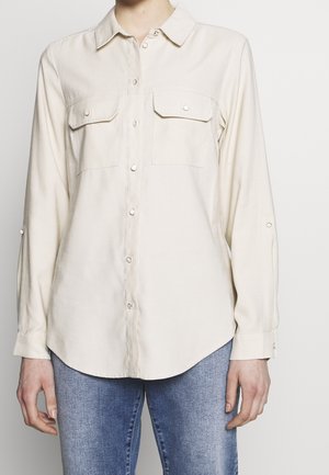 Light beige button-up shirt with a collar, two chest pockets, and roll-up sleeves. Made from a soft fabric with a smooth texture.