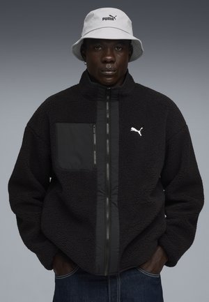 Sort fleecejakke med en glat frontpanel, lynlåslomme og hvid Puma-logo. Sammen med en lysegrå bucket hat.