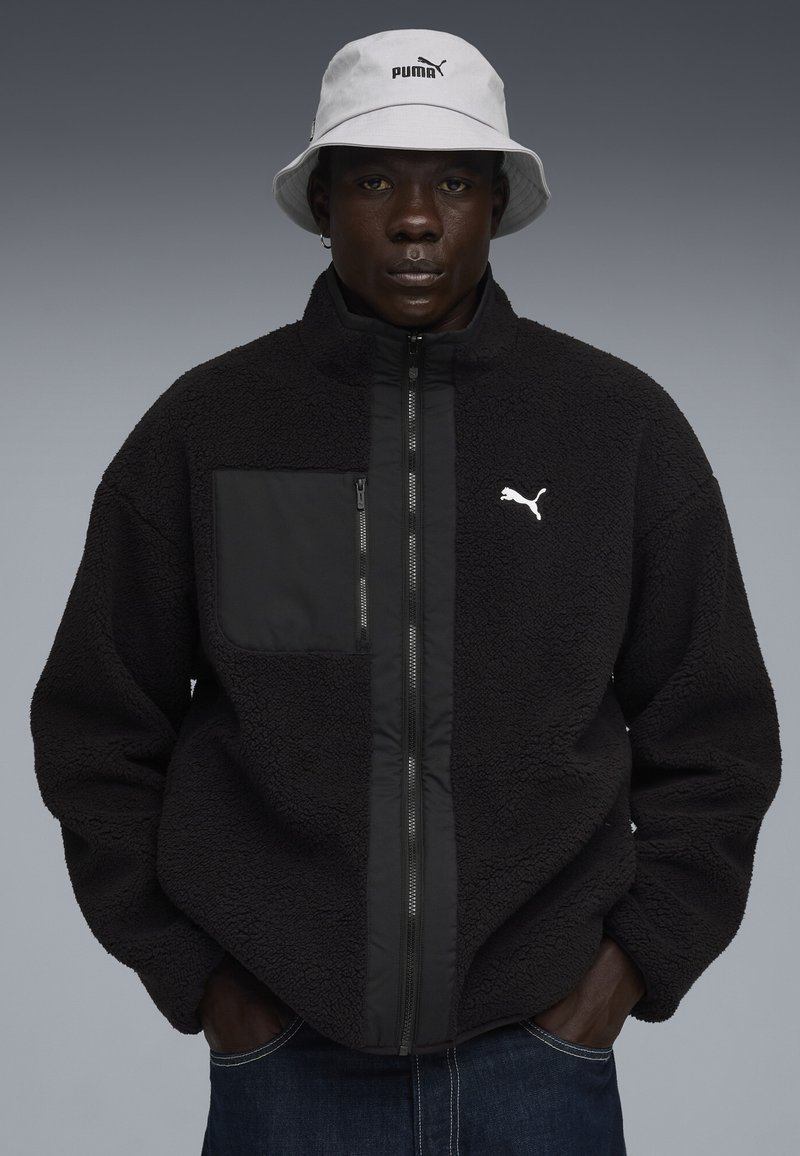 Sort fleecejakke med en glat frontpanel, lynlåslomme og hvid Puma-logo. Sammen med en lysegrå bucket hat.