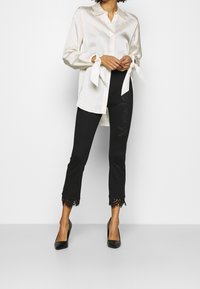 Witte satijnen blouse met strikdetails bij de mouwen, gecombineerd met zwarte cropped broek met kantaccenten aan de zoom. Zwarte hakken.