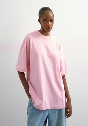 Femme aux cheveux attachés porte un t-shirt rose clair oversize et un jean bleu clair, debout devant un fond blanc uni.