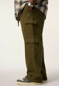 Olijfgroene cargo broek met meerdere voorzakken, gemaakt van zacht materiaal. Draagt met grijs en witte sneakers en een geruit shirt.