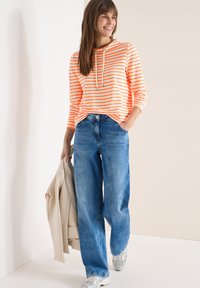 Femme portant un sweat à capuche rayé orange et blanc, jean bleu à jambes larges, baskets argentées, tenant une veste beige, souriante et marchant à l'intérieur.