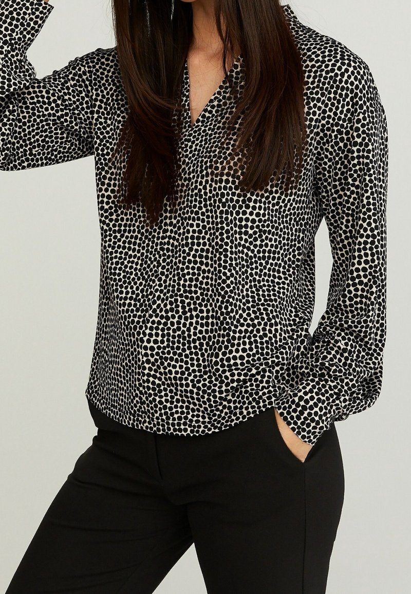 Femme portant un chemisier à pois noir et blanc à manches longues et un pantalon noir, une main dans la poche sur un fond uni.