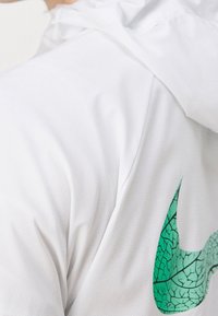 Chemise blanche à manches courtes en tissu texturé, arborant un logo Nike vert avec un motif en forme de feuille. Conception avec capuche à l'arrière.