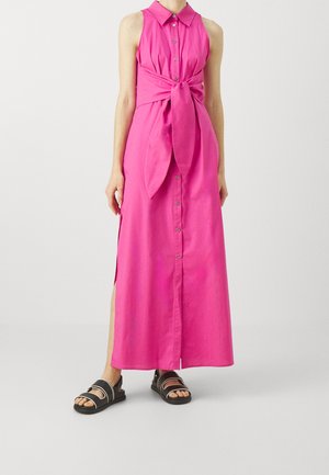 Mouwloze felroze maxi-jurk met knopen, kraag en strik aan de voorkant, gedragen met zwarte sandalen, gedragen door een persoon die voor een effen achtergrond staat.