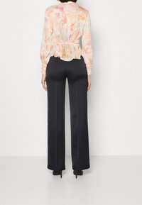 Femme portant un pantalon large noir et un blouse à manches longues florale claire, se tenant contre un fond blanc uni, de dos.