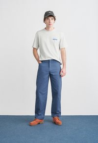 Γκρι κοντομάνικη μπλούζα με μπλε λογότυπο «DICKIES», συνδυασμένη με μπλε παντελόνια και πορτοκαλί παπούτσια. Το μοντέλο στέκεται με τα χέρια στις τσέπες.