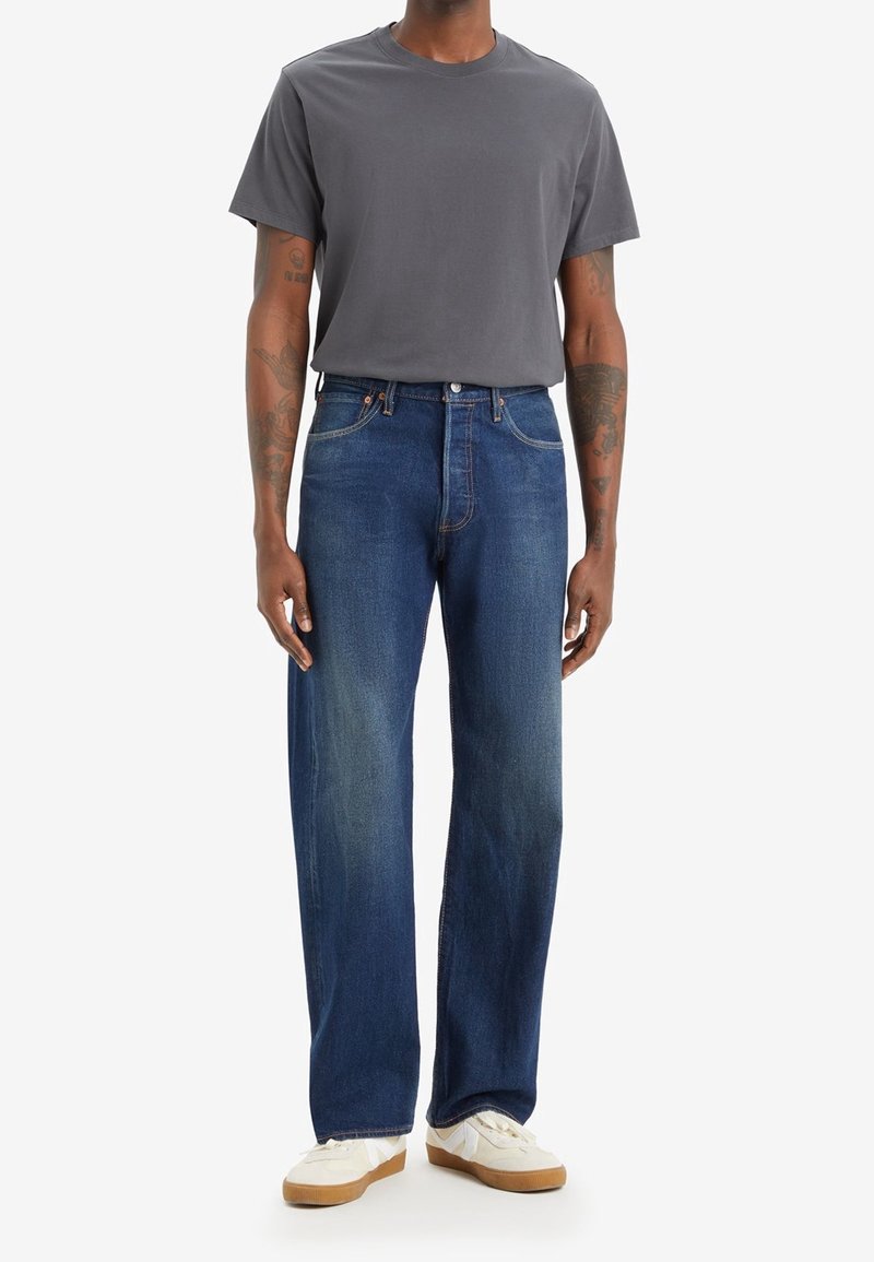 Homme debout portant un t-shirt gris foncé uni, un jean droit bleu, des baskets blanches et beiges, tatouages visibles sur les bras sur fond clair.