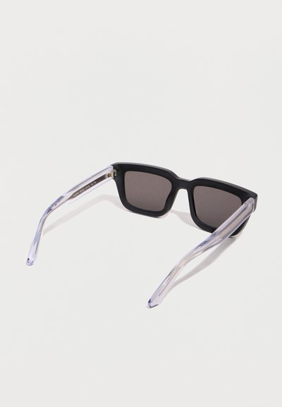 Μαύρα ορθογώνια γυαλιά ηλίου με σκούρα φακούς και διαφανή διαφανή μπράτσα, εμφανιζόμενα από μια γωνιακή πίσω όψη σε λευκή επιφάνεια.