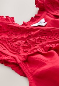 Vêtement rouge vif avec des motifs tourbillonnants brodés en relief et des bords froncés, posé à plat avec une étiquette blanche visible.
