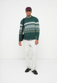 Green knitted sweater with white and yellow geometric patterns, paired with a checkered shirt, light gray pants, and black shoes. Bag slung over shoulder.