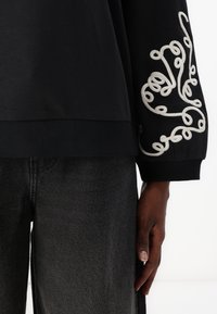 Svart hettegenser med hvite broderte svinger på ermene. Ribbestrikket kant ved nederkant og mansjetter; kombinert med mørkegrå denimbukser.