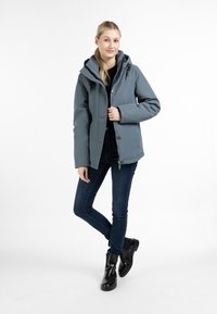 Giacca impermeabile grigia con cappuccio, chiusura con zip e bottoni, tasche sul petto. Indossata sopra una maglietta nera, abbinata a jeans scuri e stivali neri.
