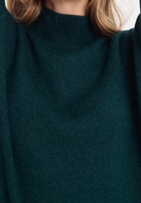 Mörkgrön stickad tröja med hög, ribbad halsringning. Mjuk textur, nedhängda axlar och lös passform för bekväm användning.