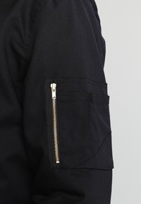Veste en tissu noir avec une poche zippée sur la manche équipée de ferrures dorées. Le matériau présente une texture lisse et une coupe ajustée.