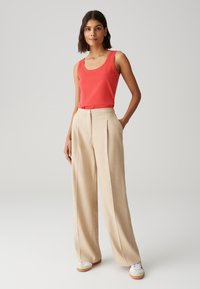 Top senza maniche rosso abbinato a pantaloni beige a gamba larga con vita alta. I pantaloni presentano delle pieghe; il modello indossa sneakers bianche casual.