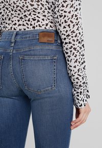 Jeans de mezclilla azul con bolsillos traseros y una etiqueta de cuero marrón que dice "DRYDEN". Una blusa con un patrón de manchas negras cubre los brazos.