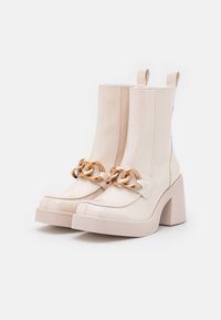 Patent beige nilkkurit, joissa on paksu korko, ja jotka sisältävät suuren kultaketjun yksityiskohtana edessä sekä vedettävyyslenkit takana.