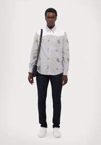 Camicia grigia con ricami floreali e colletto bianco, abbinata a jeans scuri e sneakers bianche; il modello tiene una borsa piccola.