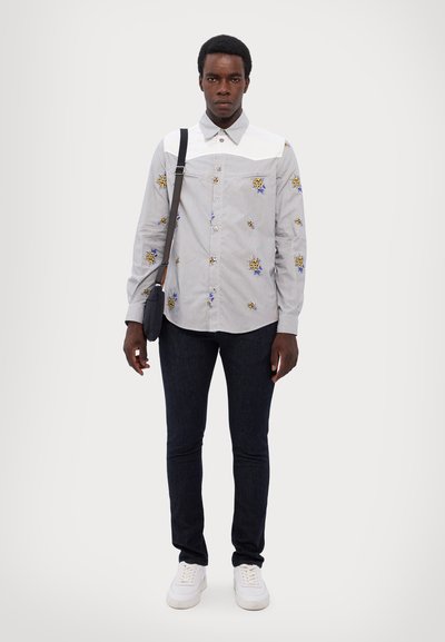 Camisa de botones con bordados florales en gris y cuello blanco, combinada con jeans oscuros y zapatillas blancas; el modelo sostiene un bolso pequeño.