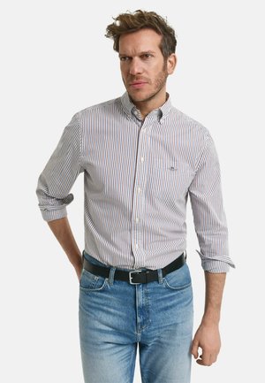 Weißes Hemd mit roten und blauen vertikalen Streifen, lange Ärmel hochgerollt, getragen mit hellblauen Jeans und einem schwarzen Gürtel.