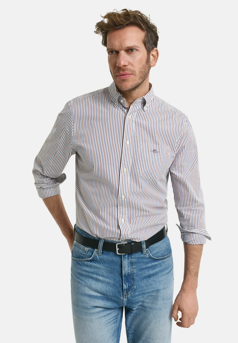 Weißes Hemd mit roten und blauen vertikalen Streifen, lange Ärmel hochgerollt, getragen mit hellblauen Jeans und einem schwarzen Gürtel.