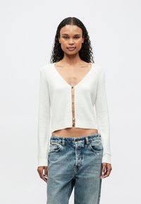 Mujer con cabello rizado que lleva un cárdigan corto blanco con botones de letras que dicen "FUTURO" y jeans azules holgados frente a un fondo sencillo.