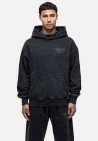 Felpa nera con cappuccio, tasca frontale, cordino e dettagli del logo sul petto. Realizzata in tessuto morbido e testurizzato. Vestibilità ampia.