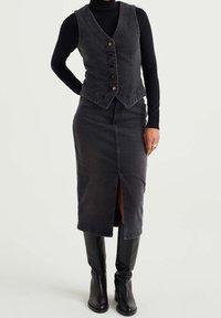 Kvinna som bär en svart polotröja, en mörkgrå knappad denimväst, en matchande blyertskjol med framklippning och svarta knä-höga stövlar.