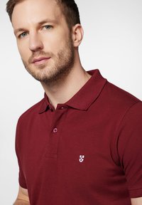 Burgundowa koszulka polo z teksturowanej tkaniny, z klasycznym kołnierzykiem, zapięciem na dwa guziki i małym białym logo na piersi.