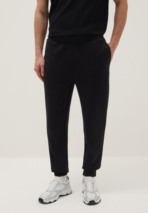 Uomo che indossa pantaloni della tuta neri aderenti con polsini alle caviglie, scarpe da ginnastica bianche e top nero senza maniche, in piedi con una mano in tasca.