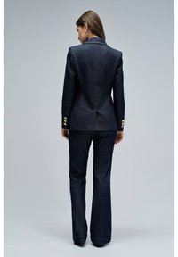 Blazer de mezclilla oscura con un diseño estructurado, ventilación trasera sencilla y botones decorativos dorados en las mangas, combinado con pantalones de mezclilla oscura de pierna acampanada.