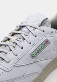 Λευκό δερμάτινο αθλητικό παπούτσι με σχέδιο κορδονιών, υφασμάτινες λεπτομέρειες, πράσινο λογότυπο Reebok και μπεζ εξωτερική σόλα. Ορατή μαλακή υφασμάτινη επένδυση.