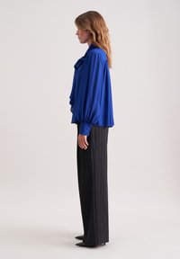 Top blouson blu con un fronte arricciato, maniche lunghe a palloncino, abbinato a pantaloni neri a righe verticali a vita alta e décolleté neri a punta.