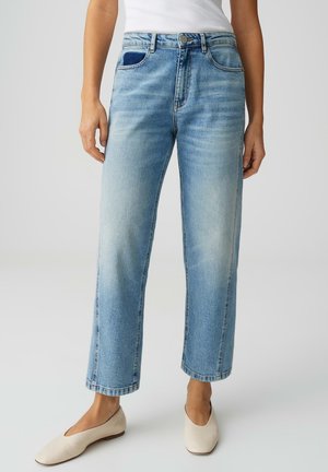 Donna che indossa jeans a gamba dritta azzurri chiaro, canottiera bianca e scarpe basse color crema, in piedi contro uno sfondo semplice.