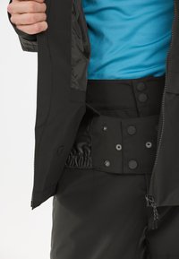 Schwarze Außenjacke mit glatter Textur, ausgestattet mit Druckknopfverschlüssen und einem elastischen Bund an der Hose; getragen über einem blauen Hemd.