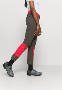 Spodnie turystyczne zwężane, ciemnobrązowe z jasnoróżowymi panelami na kolanach, bocznymi suwakami i czerwoną talią, w połączeniu z szarymi butami turystycznymi.