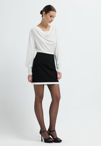 Weißes, langärmeliges Oberteil mit Drapierung, kombiniert mit einem schwarzen Minirock mit weißem Saum. Das Model trägt schwarze, durchsichtige Strumpfhosen und schwarze, geschnürte High Heels.