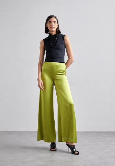 Zwarte mouwloze top gecombineerd met hoge, wijde felgroene broeken. De broek heeft een soepele afwerking en een subtiele glans. Zwarte hoge hakken.
