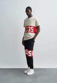 Beige t-shirt met een groot rood en wit "CDS" grafisch ontwerp over de borst, gecombineerd met zwarte broek met vergelijkbare rood en witte grafische patches.