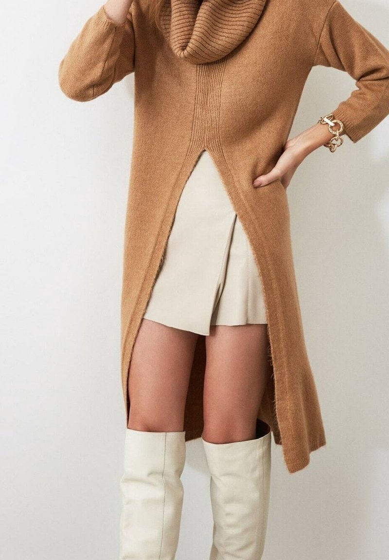 Robe-pull col bénitier de couleur camel avec une fente à l'ourlet. Confectionnée en tissu doux, elle présente des détails côtelés et est associée à une jupe plissée beige ainsi qu'à des bottes blanches montantes au-dessus du genou.