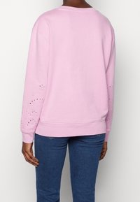 Sudadera rosa claro con cuello redondo y mangas largas, que presenta patrones florales recortados en los dobladillos de las mangas. Textura de tela suave.