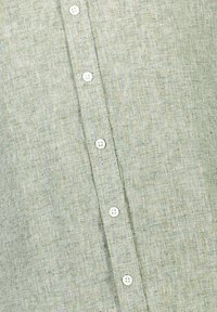 Camisa de lino verde claro con una superficie texturizada y un patrón sutil, con botones blancos en la parte delantera. Diseño de cuello simple.