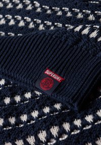 Sudadera de punto con un diseño en azul marino y crema, con un cuello de canalé y una etiqueta roja de "Superdry" cosida en el lateral.