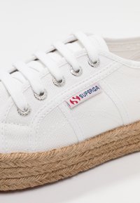 White canvas sneaker with woven jute platform sole, featuring metal eyelets and white laces. Small red and blue brand logo on side.