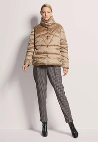 Puffermantel in hellem Beige mit hohem Kragen und Knopfverschluss. Gemusterte Hose in Schwarz und Weiß. Das Model trägt schwarze Stiefeletten.