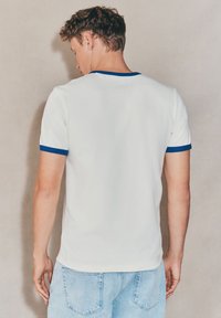 Biała koszulka z niebieskim prążkowanym kołnierzem i wykończeniami na rękawach, z okrągłym dekoltem i luźnym krojem, zestawiona z jasnoniebieskimi dżinsami.